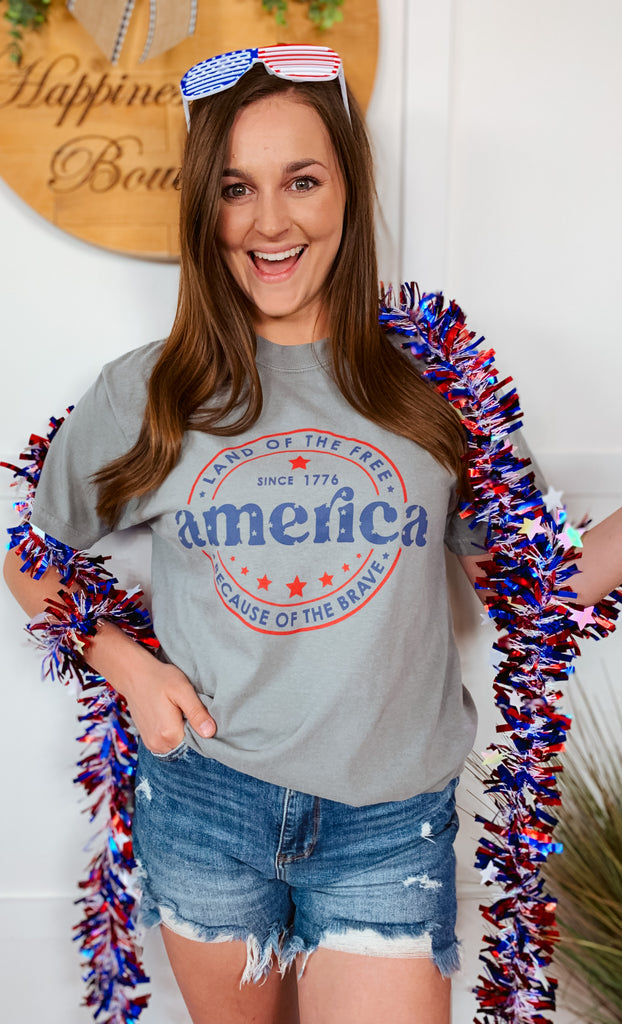 Celebrate this 4th of July in this America Land Of The Free Because Of The Brave graphic tee.  Screen printed on this grey Comfort Colors t-shirt